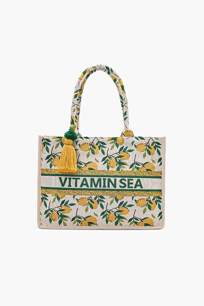 A tote bag with a yellow lemon and green leaf pattern on a cream background, featuring a green and white horizontal stripe across the front with the text 'VITAMIN SEA' and yellow tassels on the handles.